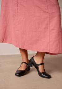 Svarta läder Mary Jane-klackar med fyrkantig tå, ett utskärningsdesign på sidorna och en låg blockklack. Bärs med en rosa linnekjol.