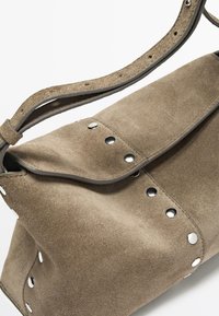 Tasche aus taupefarbenem Wildleder mit silbernen Metallnieten und verstellbarem Riemen auf weißem Hintergrund.