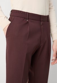 Burgundy tailored trousers with a smooth texture, pleated front, elastic waistband, and side pockets. Close-up view highlighting fit and design.