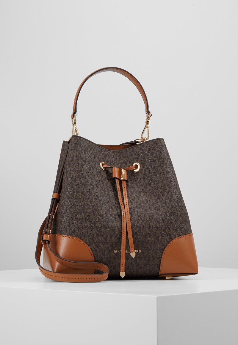 Sac seau en cuir marron et beige avec fermeture à cordon, poignée supérieure et bandoulière, placé sur un piédestal blanc sur fond gris.