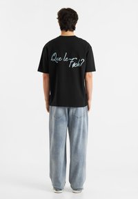 Svart bomull T-skjorte med blå tekst på baksiden som sier "Que le F**k?" Kombinert med lyseblå, vide denimjeans.