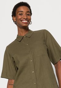 Camisa de manga curta verde-oliva com fecho de botão, gola, bolso de peito único e tecido texturizado.