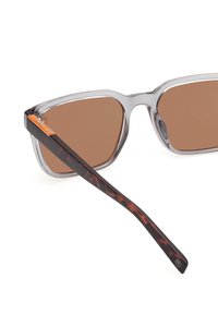 Läpinäkyvä kehysaurinkolasit ruskeilla linsseillä; teksturoitu kilpikonnakuosiset tempurit; ohuet hopeakoristeet sarveksiin lähellä saranan kohtaa.