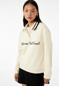 Kremowy sweatshirt z czarnym paskiem przy kołnierzu, z wyhaftowanym napisem "California West Coast" na przodzie. Miękka tekstura, luźny krój.