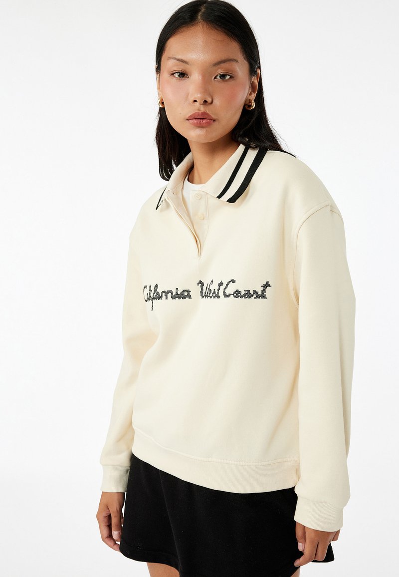 Kremowy sweatshirt z czarnym paskiem przy kołnierzu, z wyhaftowanym napisem "California West Coast" na przodzie. Miękka tekstura, luźny krój.