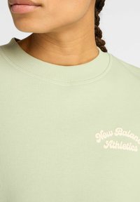 Gros plan d'une personne portant un sweat-shirt vert clair avec le texte blanc "New Balance Athletics", montrant le cou et des cheveux tressés.