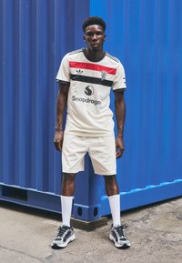 Jersey da uomo color crema con strisce rosse e nere, dotato di logo e branding. Indossato con pantaloni corti chiari e sneakers nere, su uno sfondo blu.