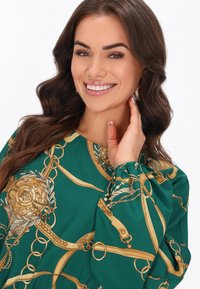 Hosszú ujjú zöld blúz aranylánccal és díszítő mintával. Az összegyűrt ujjak textúrát és részletességet kölcsönöznek a dizájnnak.
