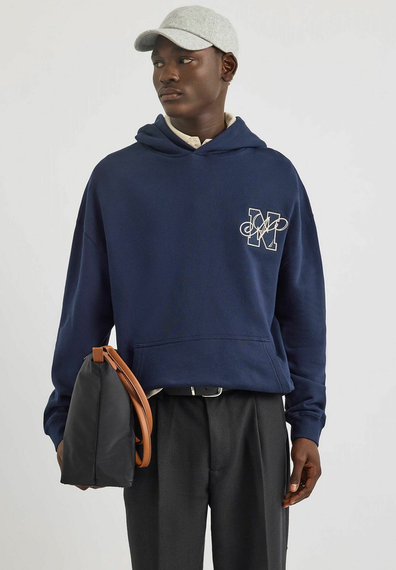 Felpa blu navy con tasca frontale e logo ricamato. Abbinata a pantaloni neri e una borsa nera con tracolla marrone. Cappello grigio.