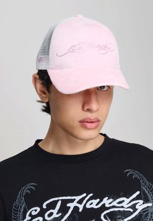 Młody mężczyzna noszący jasnoróżową siatkowaną czapkę typu trucker z napisem z kryształków i czarną koszulkę Ed Hardy z białymi i szarymi wzorami.
