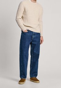 Cremfarbener Strickpullover mit einem strukturierten Muster, kombiniert mit dunkelblauen Jeans. Hellbraune Schuhe runden das Outfit ab. Schlichtes und lässiges Design.