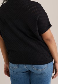 Osoba v černém krátkém pleteném topu s texturou a modrých džínech, viděná zezadu na neutrálním pozadí.