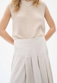 Beige stickad ärmlös topp med v-ringning, tillsammans med en ljusgrå, veckad linnekjol. Fokusera på textur och neutrala färger.