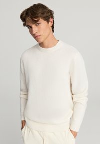 Sweter w kolorze kremowym, wykonany na drutach, z okrągłym dekoltem, prążkowanymi mankietami i dolnym brzegiem. Wykonany z miękkiego, teksturowanego materiału. Noszony z jasnymi spodniami.