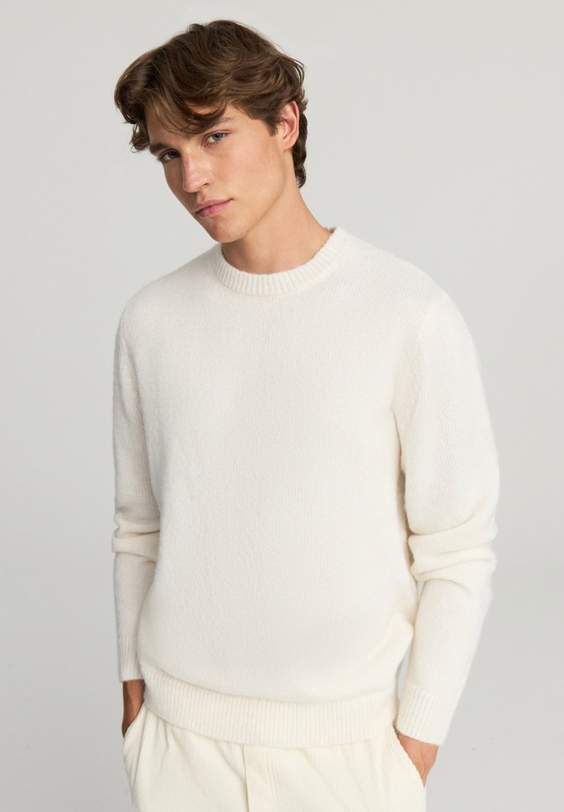 Sweter w kolorze kremowym, wykonany na drutach, z okrągłym dekoltem, prążkowanymi mankietami i dolnym brzegiem. Wykonany z miękkiego, teksturowanego materiału. Noszony z jasnymi spodniami.