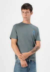 Yng mand med kort mørkt hår iført en grå t-shirt med en blå grafik på brystet og blå jeans, der viser en detaljeret tatovering på underarmen.