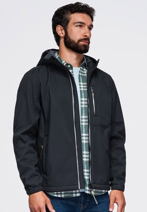 Homme avec barbe portant une veste noire à capuche par-dessus une chemise écossaise verte et blanche et un jean bleu, regardant vers la droite.