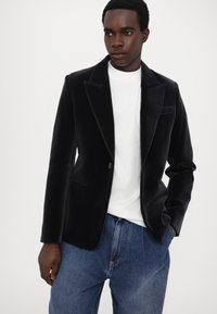 Juodas aksominis švarkas su vienu mygtuku, notch tipo apykakle ir šoninėmis kišenėmis, derinamas su balta marškinėliais ir mėlynomis džinsinėmis kelnėmis.