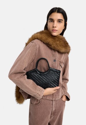 Kobieta w brązowej kurtce z futrzanym kołnierzem, trzymająca małą czarną plecioną torebkę, stojąca na jednolitym jasnym tle.