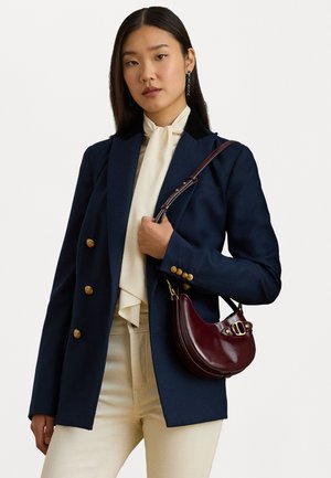 Kék blézer arany gombokkal, fekete gallérral és krémszínű nyakkendővel; burgundi bőr kézitáskával kombinálva; a modellen krémszínű nadrág van.