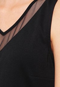 Svart ärmlös topp med en djup V-ringning och en genomskinlig mesh-overlay. Slät textur med en subtil glans och en liten sidoficka.