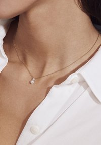 Guld kædeda necklace med en rund perle og en lille diamant accent, båret over en hvid skjorte med krave. Enkel og elegant design.
