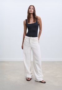 Svart bodysuit med fyrkantig halsringning, ihopparad med löst sittande, högmidjade vita jeans och bruna sandaler på en neutral bakgrund.