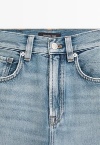 Světle modré džíny s vysokým pasem, mají kovový knoflík, přední zip a design s pěti kapsami. Značka je viditelná na pase.