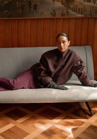 Oversized dark brown wool coat with high collar, paired with maroon trousers and black leather gloves, lounging on a light gray sofa.