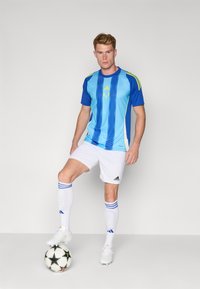 Blå og lyseblå stribet fodboldtrøje med gule accenter, hvide shorts og hvide sokker, stående på en fodbold med en fokuseret stilling.