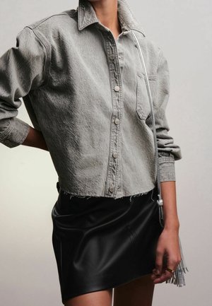 Personne portant une chemise en jean grise boutonnée avec des manches retroussées et une jupe en cuir noire, tenant un sac à main argenté avec des franges.