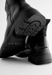 Bottines en cuir noir avec une semelle épaisse, présentant un motif de crantage texturé et une finition lisse, complétées par un talon renforcé.