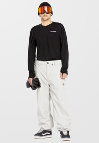 Chemise noire à manches longues, pantalon de neige blanc, gants noirs et lunettes de ski réfléchissantes. Bonnet gris, baskets noires avec semelle blanche.