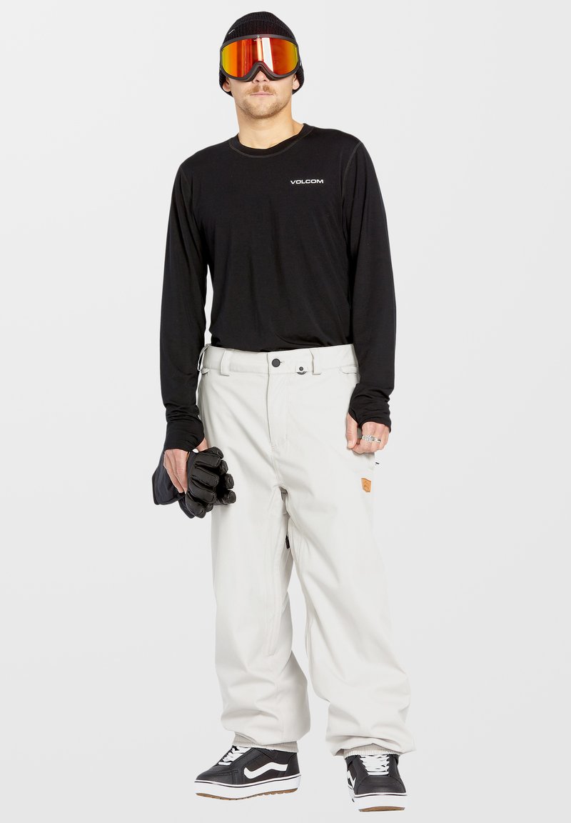 Chemise noire à manches longues, pantalon de neige blanc, gants noirs et lunettes de ski réfléchissantes. Bonnet gris, baskets noires avec semelle blanche.