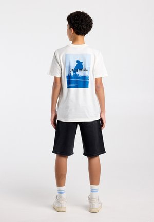 Jong persoon met kort krullend haar die een wit T-shirt draagt met blauwe California skateboardprint, zwarte korte broek, witte sneakers en gestreepte sokken.