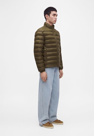 Jeune homme debout portant une doudoune vert olive, un jean large bleu clair et des baskets marron sur un fond blanc.
