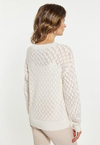 Lekki sweter z dzianiny w kolorze ecru z diamentowym wzorem koronkowym, z prążkowanymi mankietami i dołem. Długie rękawy, z tyłu widoczny jest otwarty wzór tkaniny.