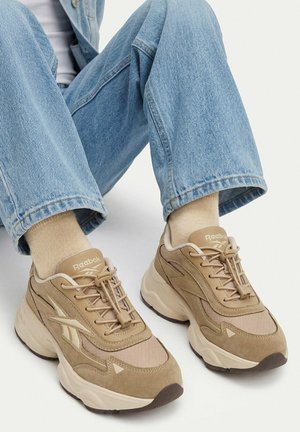 Persoon draagt beige Reebok sneakers met dikke zolen, lichtbeige sokken en lichtblauwe spijkerbroek, zittend met gebogen benen.