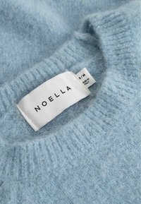 Hellblauer, flauschiger Strickpullover mit einem weißen Stoffetikett, das den Markennamen "NOELLA" und die Größe "S/M" zeigt, hergestellt in Italien.