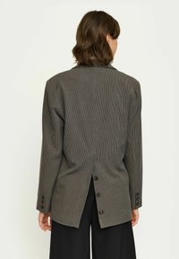 Blazer grigio a quadretti con uno spacco sul retro e chiusura a bottoni. Presenta maniche lunghe e un design su misura. Il materiale appare strutturato.