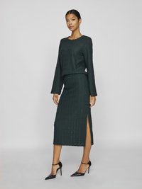 Σέτ από πλεκτό κοντό τοπ και φούστα με ψηλή σχισμή σε σκούρο πράσινο, με υφή και μακριά μανίκια, συνδυασμένα με μαύρες μυτερές γόβες.