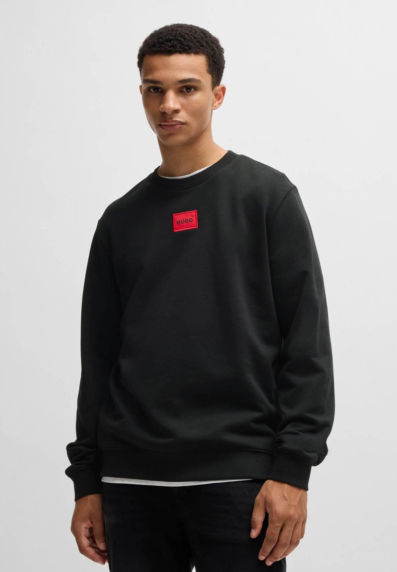 Svart sweatshirt i bomull, med rund hals och lång ärm. Röd rektangulär lapp med "HUGO"-text på framsidan.