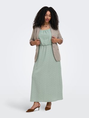 Kvinne med krøllete hår iført en lysegrønn prikkete maxikjole, beige blazer, brune hæler og et halskjede mot hvit bakgrunn.