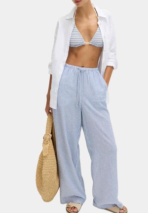Donna che indossa pantaloni larghi a righe blu e bianche, top bikini blu a motivo, camicia bianca aperta, sandali beige, che tiene una grande borsa intrecciata.