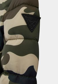 Giacca puffer mimetica con ampi motivi verdi e beige, dotata di una toppa in gomma nera a forma triangolare sulla manica superiore.