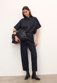 Mörk denimskjorta med kort ärm, knappstängning och kontrasterande sömmar, matchad med mörka jeans och svarta läderstövletter. Håller en svart väska.