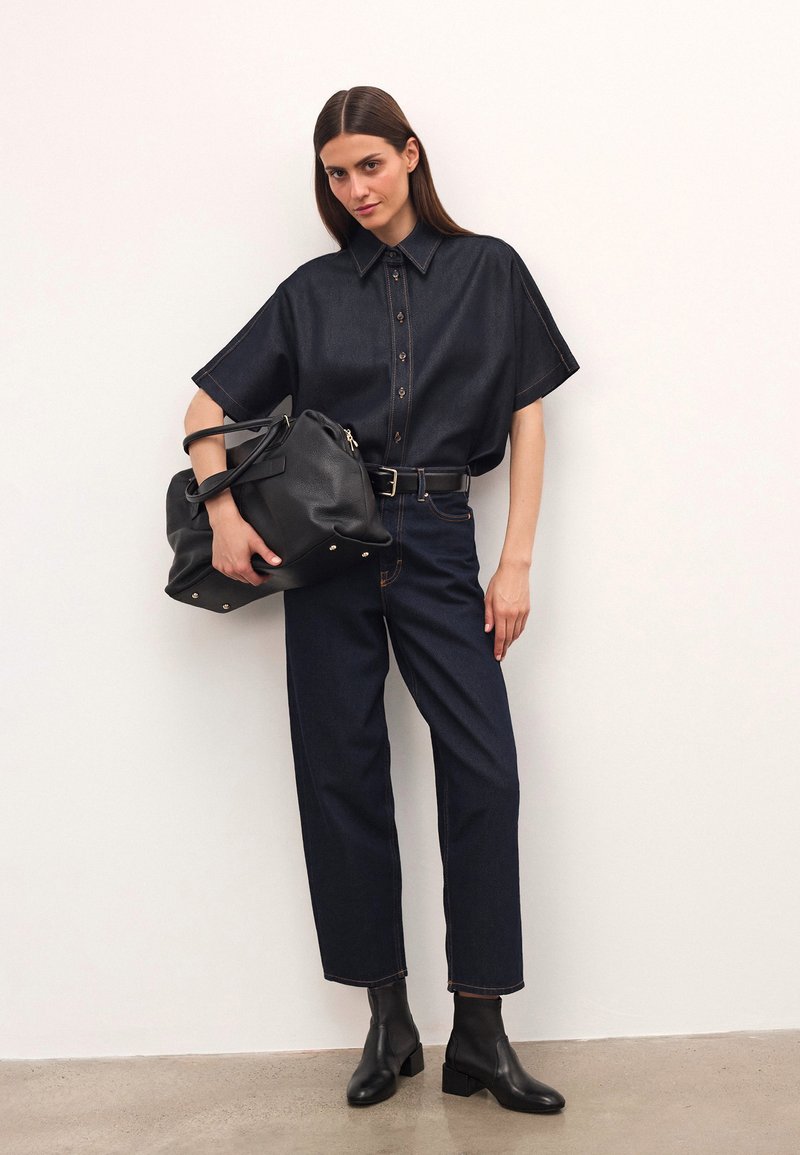 Mörk denimskjorta med kort ärm, knappstängning och kontrasterande sömmar, matchad med mörka jeans och svarta läderstövletter. Håller en svart väska.