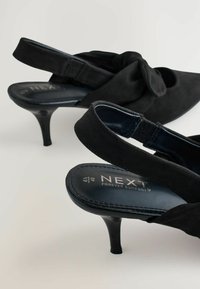 Schwarze Wildleder-Pumps mit Slingback-Design, die an der Vorderseite mit einem Knotenakzent versehen sind. Die Innensohle bietet gepolsterten Halt und ist mit "NEXT FOREVER COMFORT" gekennzeichnet.