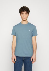 Short-sleeved, crew neck t-shirt in light blue cotton with a subtle logo on the chest. Styled with dark blue jeans, plain texture.