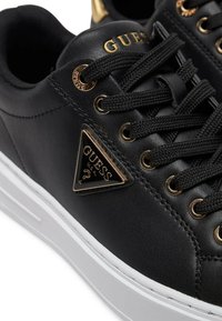 Baskets en cuir noir avec une semelle en caoutchouc blanche, présentant des œillets dorés, des lacets noirs et un accent de logo doré triangulaire sur le côté.